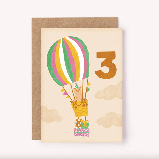 Featuring an illustrated bear in a striped hot air balloon, carrying a stack of three wrapped gifts. There is a bold hand-lettered number 3 on a soft beige background. The subtle colour palette makes it a gender neutral greeting card, ideal for celebrating any child turning three.